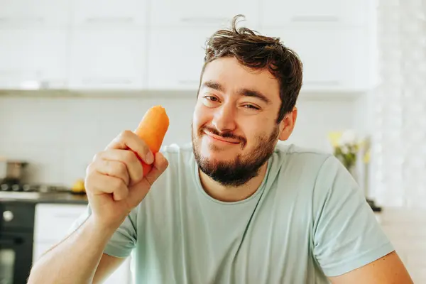 stock image Smiling derisively at the camera, a young man holds a raw orange carrot in his hand. Maybe he is on a diet to improve his health. The guy does not want to eat it.