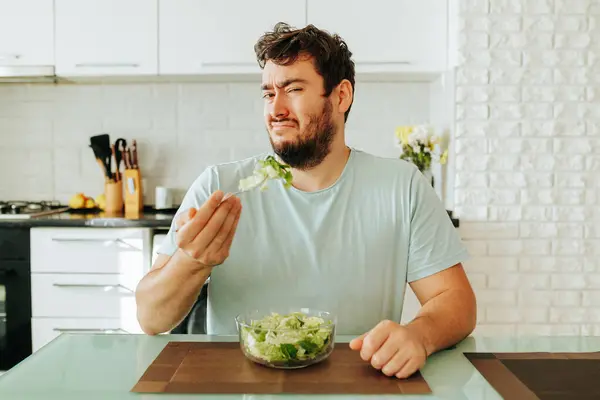 stock image Frustrated by the need to eat unloved food, a young guy holds a fork with a green salad in his hand. Diet food is not what the guy dreamed of. Stop diet, healthy food, vegan food.
