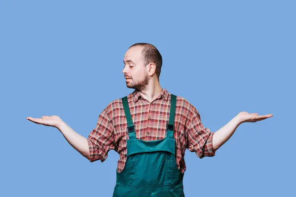 stock image side view of An expressive man captured with his hands outstretched in a curious and inquisitive pose, dressed in a coverall, against a blue studio background.