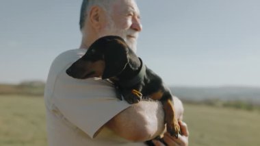 Gün batımında bir tarlanın dingin atmosferinde yaşlı bir adam sevgili dachshund 'unu beşikte tutarak arkadaşlığın güzelliğini gösterir..