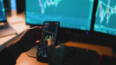 Ellerinin akıllı telefon ekranında yazı yazarken ve hesap makinesi kullanırken çekilmiş yakın çekim görüntüsüyle, monitörde gösterilen grafikleri ve grafikleri titizlikle analiz ediyor. Bu fotoğraf yoğun bir odak yayar.
