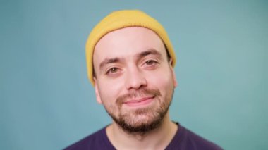 Sarı şapkalı bir adam, yüzünde geniş ve ışıl ışıl bir gülümsemeyle, neşe ve memnuniyet dolu bir hava yayıyor, bir stüdyoda, mavi bir arkaplana karşı, fotokopi aletiyle kameraya dikkatlice bakıyor..