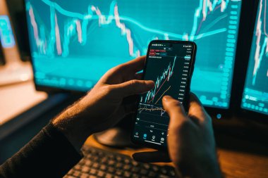 Ellerinin akıllı telefon ekranında yazı yazarken ve hesap makinesi kullanırken çekilmiş yakın çekim görüntüsüyle, monitörde gösterilen grafikleri ve grafikleri titizlikle analiz ediyor. Bu fotoğraf yoğun bir odak yayar.