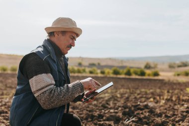 Yaşlı bir çiftçi, toprak koşullarını inceleyen dijital bir tabletle hasadı takip ediyor. Bu görüntü, tarımda teknolojiyle karşılaşma deneyiminin özünü yakalar..