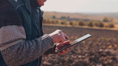 Hasatın ortasında, yaşlı bir çiftçi elleri tarım arazilerini incelemek için dijital tablet kullanır. Gelenek, mahsul sağlığı ve verimliliği güvence altına aldığı için teknolojiyle buluşuyor. Kapat.