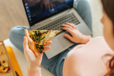 Dizüstü bilgisayarında yazı yazan bir kadının bir dilim pizza tutarken çekilmiş yakın plan fotoğrafı. Genç bir kadın evdeki kanepesinde oturmuş online alışveriş sitelerine bakarken bir dilim pizzanın tadını çıkarıyor.