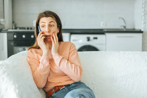 stock image A young woman is sitting on her sofa at home, talking on her smartphone, with a puzzled expression on her face as she tries to understand what the person on the other end of the line is saying.