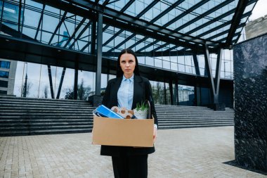 White Collar Worker Holding Cardboard A Sign of Joblessness. An unemployed woman holds a cardboard box, looking worried and upset after experiencing workloss and loss work. clipart