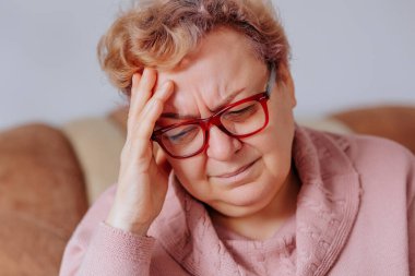 Oturma odasında şiddetli bir baş ağrısı ve baş ağrısı yüzünden elleriyle başını tutan kilolu yaşlı bir kadın migren ağrısı çekiyor.