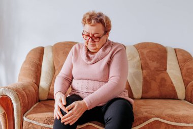 Yaşlı bir kadının evdeki bacak ağrısıyla mücadelesinin ağırlığı, evdeki koltukta oturan şişman bir kadın, bacak ağrısı ve obezite ve yaşlanma stresi yüzünden üzgün ve üzgün görünüyor..