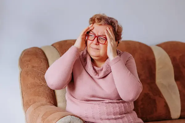 stock image Senior woman with a throbbing headache, seeking relief by placing her hands on her forehead in her cozy living room, Living with Pain Overweight Senior Woman Struggles with Aches