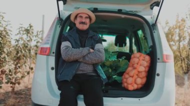 Hasat zamanındaki yaşlı çiftçi arabanın bagajında, patates çuvallarıyla çevrili, devam eden hasat sezonunu düşünceli bir şekilde sunuyor..