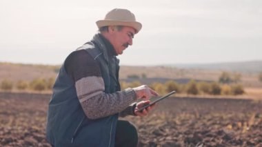 Deneyimli kıdemli bir çiftçi, bıyıklı, hasat sırasında dijital tablet kullanıyor. Uzmanlığı eski bilgeliği modern teknolojiyle harmanlıyor, ekin koşullarını optimize ediyor..