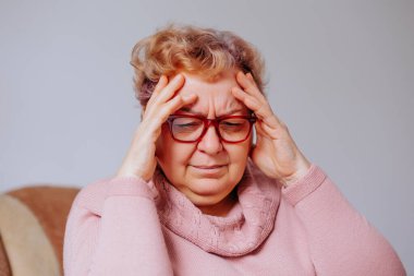 Aşırı kilolu yaşlı kadın migren oluyor, elleri başının üstünde, evinde huzur ararken, Baş Ağrısının Üstesinden gelen Yaşlı Kadın Evde Yardım Buldu