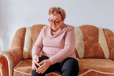 Yaşlanmış bir kadının evde bacak ağrısıyla mücadele etmesi, evdeki koltukta oturan şişman bir kadının bacak ağrısı ve obezite ve yaşlanma stresi yüzünden üzgün ve üzgün görünmesi..