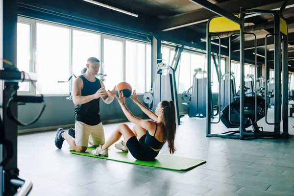 stock image An american man and a caucasian woman, with their personal trainer's assistance, performing functional exercises using a training ball to enhance their physical fitness.