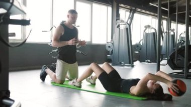 Amerikalı bir erkek ve beyaz bir kadın, kişisel antrenör yardımlarıyla, fiziksel uygunluklarını arttırmak için bir eğitim topu kullanarak egzersiz yapıyorlar..