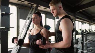 Spor salonunda, destekleyici bir erkek antrenör bir kadına bir dambılı kaldırırken rehberlik eder. Atletik Destek Erkek Kişisel Eğitmen Halter Kadın YardımcısıName