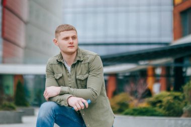 Bu güzel açık hava fotoğrafında, sarışın yakışıklı genç bir adam doğrudan kameraya bakıyor. Yalnız ve göz kamaştırıcı Çevrede Genç Bir Adam