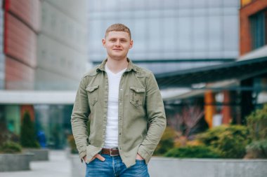 Mutlu ve tatmin olmuş sarışın bir adam bu portrede, yüzünde neşeli bir gülümsemeyle doğrudan kameraya bakarken görülüyor. Açık hava kentsel ortamının gençlik ve modaya uygun havası genel olarak