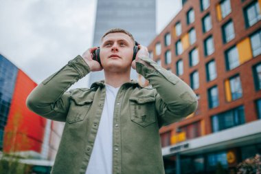 Şapka ve kulaklık takan genç bir adam, arka plandaki şehrin güzel manzarasının tadını çıkarıyor. Açık Hava Melodileri Genç Erkek Şehirdeki Müziğinin tadını çıkarıyor