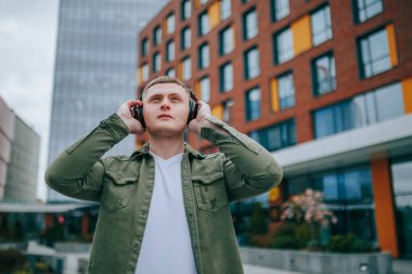 Siyah tişörtlü, kulaklık takan ve arkasındaki yüksek binaların manzarasından hoşlanan genç bir adam. Açık hava Harmony Genç Erkek Kulaklıkla Rahatlıyor