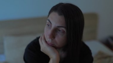 Genç bir kadın sessizce yatağında oturuyor, yatak odasının karanlığına bakıyor. Yüzündeki ifade, depresyonla mücadelesine eşlik eden derin üzüntü ve yalnızlığı ortaya çıkarıyor..