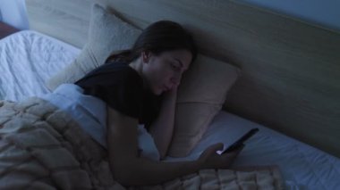 Bir kadın gece yatağında uzanıyor, akıllı telefonuna dalmış. Karanlık oda uykusuzluk sorununu yansıtıyor, çünkü uyumakta zorlanıyor..