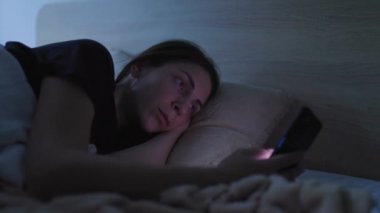 Uykusuz bir kadın gece yatağında uzanmış, akıllı telefonuna kafayı takmış. Karanlık yatak odası uykusuzluk ve uyku endişesi ile mücadelesini vurguluyor..