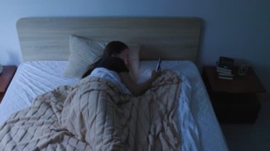 Bir kadın gece yatakta uzanıyor, akıllı telefonu karanlık odayı aydınlatıyor. Uykusuzluğu ve telefon bağımlılığı uykusuzlukla boğuşurken belli oluyor..