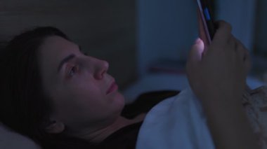 Uykusuz bir kadın gece yatağında, akıllı telefonunu kullanıyor. Odadaki loş ışık uykusuzluk ve telefon bağımlılığı sorununu vurguluyor..