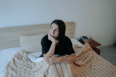 Genç bir kadın yatağının kenarında oturur, başucundaki lambanın yumuşak parıltısıyla yıkanır. Yüzünde derin bir üzüntü var. Yalnızlık ve depresyonun ağır yükünü yansıtıyor..