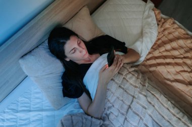Geceleri yatakta uzanan bir kadın akıllı telefonuna gömülür. Karanlık oda uykusuzluğunu ve uyumakta zorlandığını gösteriyor..