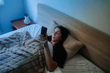 Uykusuz bir kadın gece yatağında, akıllı telefonunu kullanıyor. Odadaki loş ışık uykusuzluk ve telefon bağımlılığı sorununu vurguluyor..