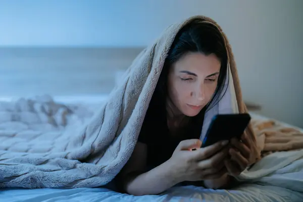 Bir kadın gece yatağında, akıllı telefonuna dalmış bir şekilde uzanıyor. Karanlık oda onun uykusuzluk ve mobil cihazına olan bağımlılığını vurguluyor..