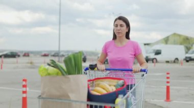 Bir kadın, çeşitli ürünlerle dolu bir alışveriş arabasını süpermarketin otoparkında taşıyor. Sahne, başarılı bir alışveriş gezisinden sonra arabasına dönerken onu yakalıyor.