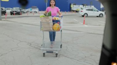 Bir kadın bir süpermarketin otoparkında içi ürünlerle dolu bir alışveriş arabasını taşırken görülüyor. Açık hava sahnesi, alışverişini tamamlayıp, alışverişini arabasına yüklemeye hazır olduğunu gösteriyor.