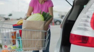 Süpermarketin otoparkında bir kadın, içi ürünlerle dolu bir alışveriş arabasını taşırken görülüyor..