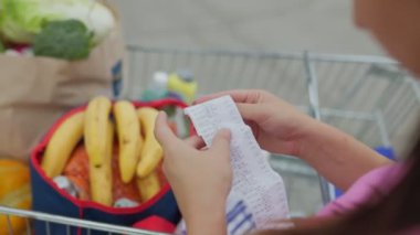 Bir kadının elinde alışveriş makbuzu, arka planda tam dolu bir alışveriş arabasıyla bir süpermarketin otoparkında görüldüğü yakın plan bir fotoğraf..