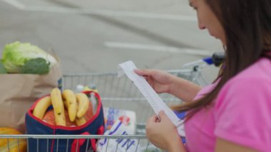 Bir kadın elinin alışveriş fişini incelerken yakın plan görüntüsü, arka planda alışveriş arabasıyla, süpermarketin otoparkında çekilmiş..