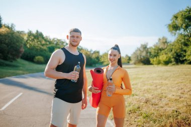 İki sporcu, bir erkek ve bir kadın, spor kıyafetleri içinde, sohbet ediyorlar ve egzersiz rutinlerinden sonra dışarı çıkıyorlar. Adamın elinde su şişesi, kadının elinde de spor paspası var..