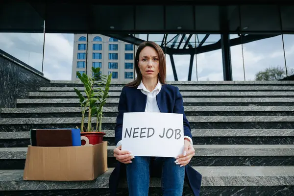 Stock image A disappointed woman sitting on the stairs, holding a carton with the inscription Job Loss on a banner, conveying her worried and unhappy emotions due to the loss of her job.