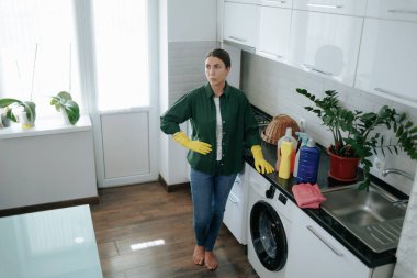 Mutfakta yorgun bir ev hanımı, sarı eldivenler giyiyor ve ev temizliğine çok ihtiyaç duyulan bir mola veriyor, yorgunluk belirtileri gösteriyor..