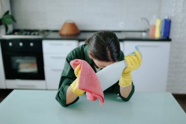 Sarı koruyucu eldivenli yorgun genç bir kadın mutfak masasında oturuyor. Yorgunluk ve hüsranla dolu bir ifadeyle onu bekleyen bitmek bilmeyen görevleri düşünüyor..