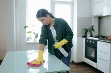 Mutfakta eldiven giyen genç bir kadın, düzenli bir şekilde masayı yıkıyor, düzenli ve temiz bir ev sahibi olma çabalarını temsil ediyor..