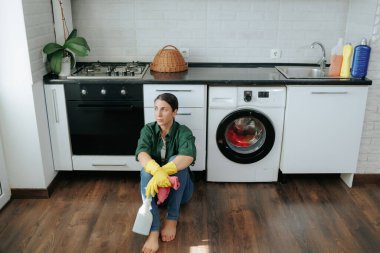 Yorucu bir temizlik seansından sonra mutfak zemininde oturan sarı eldivenli genç bir kadının otantik tasviri. Sahne onun yorgunluğunu ve hayal kırıklığını yakalıyor..