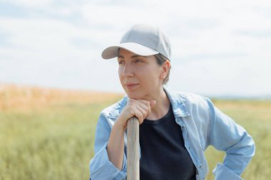 Neşeli bir dişi çiftçi, güneşli bir mısır tarlasının ortasında duruyor. Kameraya gülümserken şapkası hafifçe yana yatmış. Mısırın ve mavi gökyüzünün parlak renkleri canlı bir hava yaratır.,