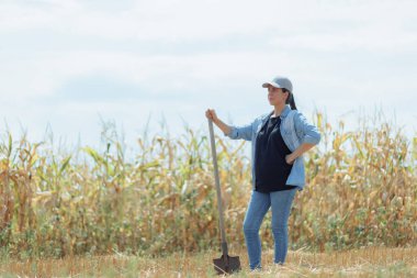 Başında şapkası ve sıradan bir kot gömleği olan bir kadın çiftçi, güneşli bir mısır tarlasının ortasında güvenle duruyor. Parlak güneş ışığı ve geniş tarım manzarası