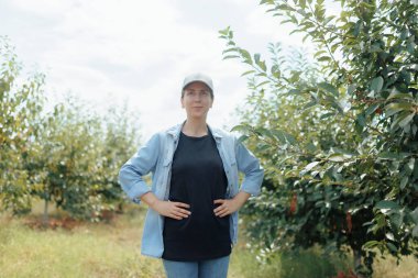 Bir meyve bahçesinde, kafasında şapka olan genç bir dişi çiftçi gelişmekte olan bitkileri inceliyor. Çevresindeki kırsal alan tarımsal çalışmalarının önemini vurguluyor..