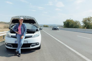 Yoğun bir otoyolda ani bir araba arızasıyla uğraşırken genç bir kadın gözle görülür bir şekilde stresli ve üzgün görünüyor. Başarısızlığın neden olduğu bozulma ve gecikmeyi göz önünde bulundurarak endişesi büyüyor..